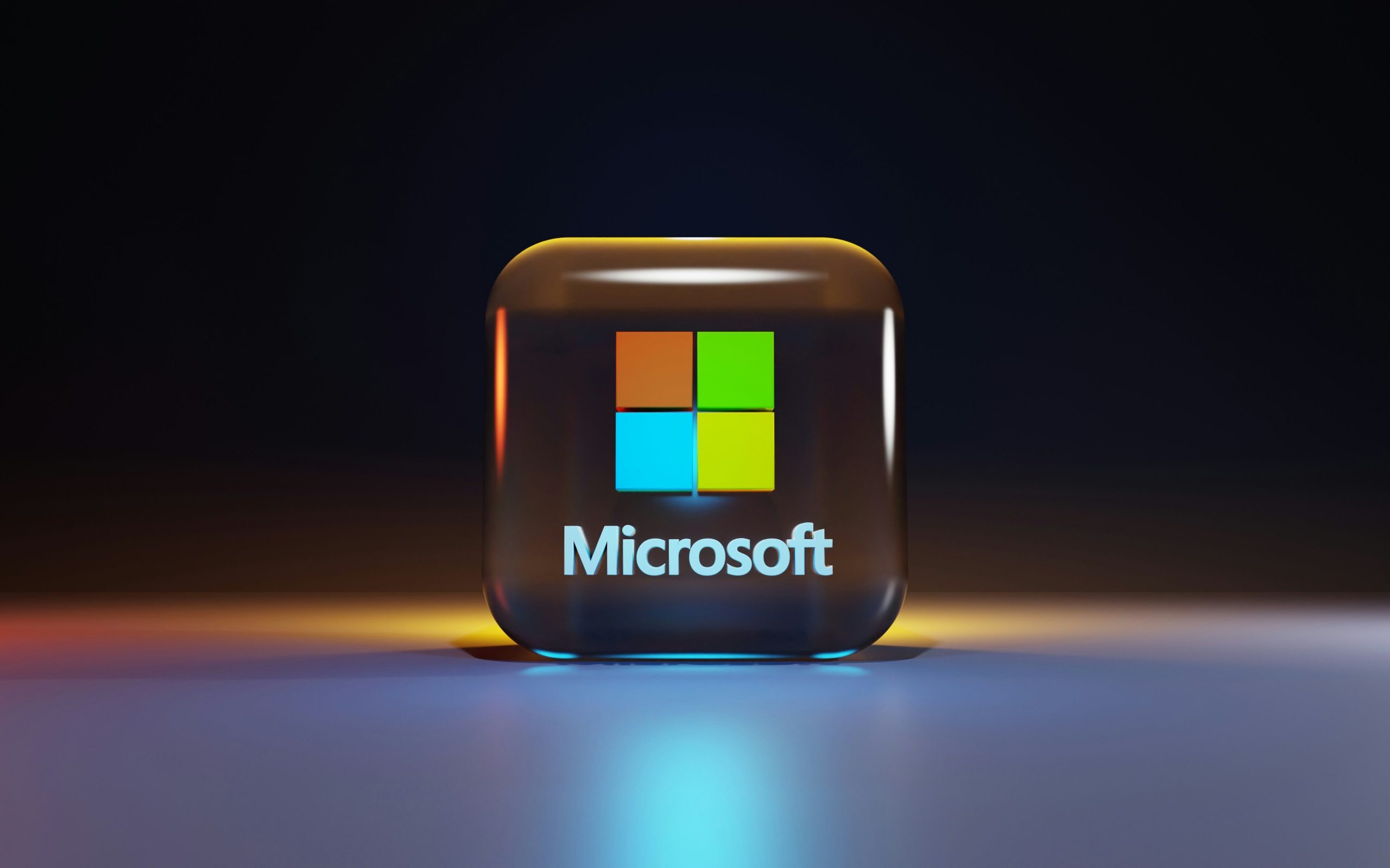 A 3D glass-like cube displaying the Microsoft logo with its four colored squares and white text, illuminated by soft blue and yellow lighting against a dark background.