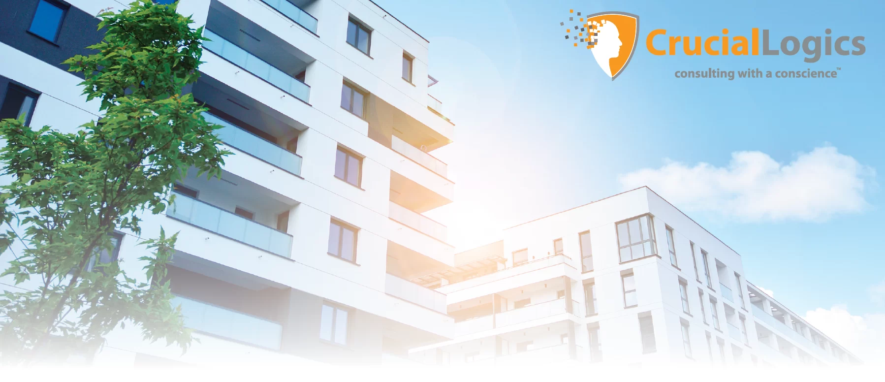 An image showing modern white apartment buildings under a bright blue sky with a few clouds; a green tree stands in the foreground; the CrucialLogics logo with the tagline consulting with a conscience appears in the upper right corner.