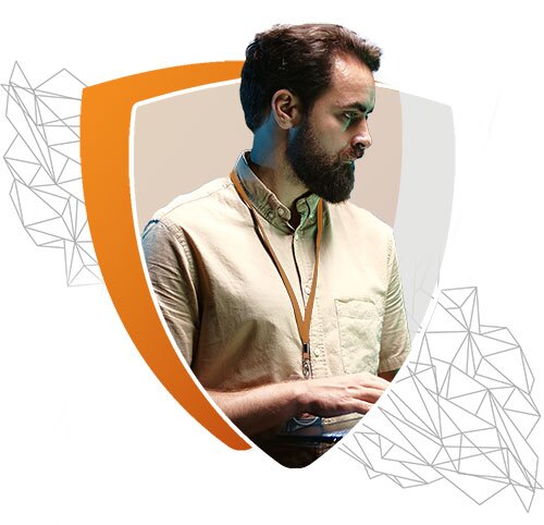 An image of a man with a beard wearing a beige shirt and lanyard, working on a laptop. The background features an orange and gray shield shape with abstract geometric line designs, symbolizing cybersecurity or IT protection.