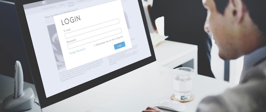 A person sits at a desk viewing a computer screen displaying a login form with fields for email and password. The workspace includes a glass of water and office materials, emphasizing secure access or authentication.