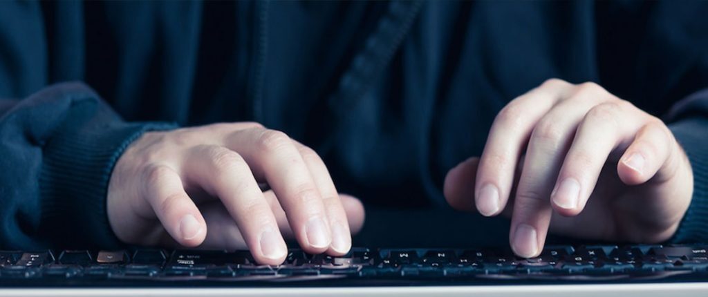A person wearing a dark hoodie types on a keyboard in a dimly lit setting. The close-up view emphasizes the hands, symbolizing coding, cybersecurity, or digital activity.