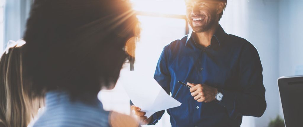A man in a blue shirt smiles while holding papers and speaking to colleagues in a bright office. The sunlight streaming in adds a warm, positive atmosphere, suggesting a productive team discussion.