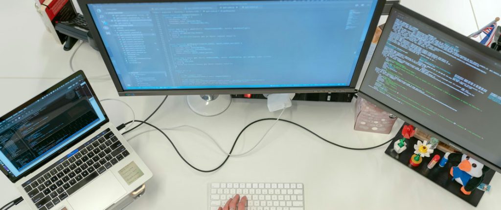 A person works at a desk with a laptop and two monitors displaying lines of code. The workspace is organized with small decorative items, indicating a software development or programming environment.