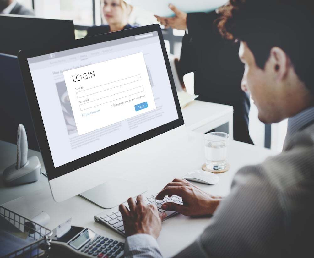 Man working at desk – computer monitor displays login page with email and password fields; office environment visible in background with colleagues conversing; glass of water and mouse beside keyboard; scene depicts workplace digital access or secure system login.