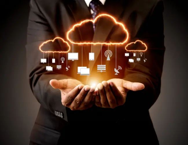A person in a business suit holds out their hands with glowing digital cloud icons above them; connected to symbols of devices and networks, representing cloud computing and data storage technology.