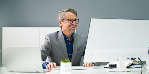 Man in a gray suit and glasses sitting at a white desk working on a desktop computer with a laptop beside him. The setting appears to be a modern office with a minimalist design, and a small potted plant adds a touch of decor to the workspace.