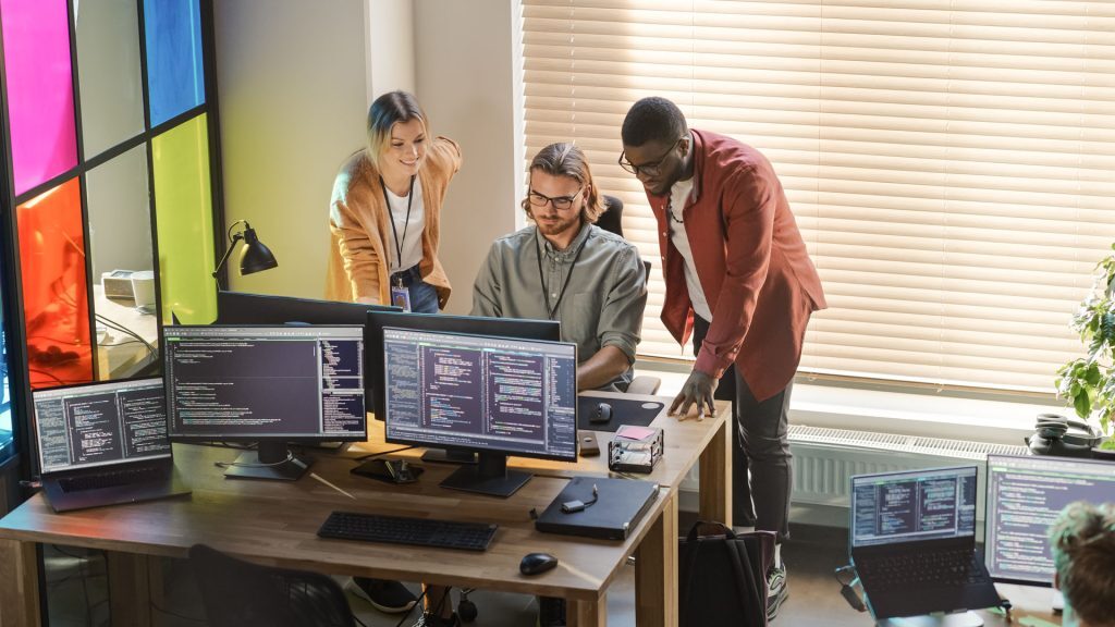 Three people collaborate in a modern office, gathered around a desk with multiple monitors displaying code. One person sits typing while the others stand beside him discussing the project. The workspace is well-lit with natural light coming through blinds and colorful glass panels in the background.