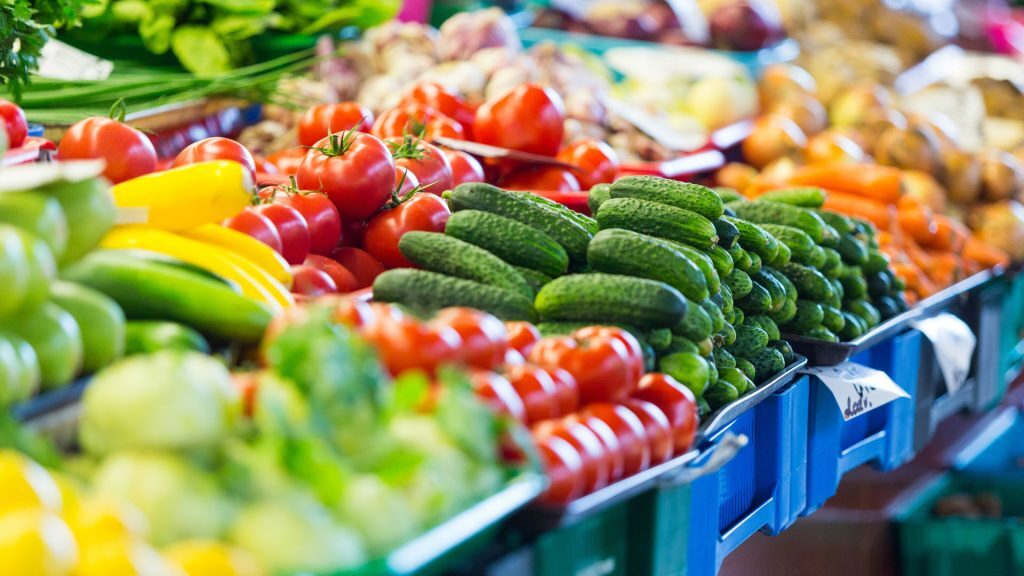 A colorful display of fresh vegetables at a market, including cucumbers, tomatoes, bell peppers, and other produce arranged in blue crates on a vendor’s stall.