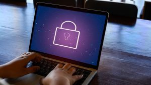 This image shows a person typing on a laptop with a digital lock icon displayed on the screen. The lock symbol, set against a purple background with a hexagonal pattern, represents cybersecurity or data protection.
