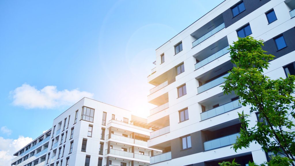 This image shows modern apartment buildings with clean, contemporary architecture. The structures have white and gray facades, large windows, and glass balconies. The photo is taken from a low angle, with sunlight creating a bright, vibrant atmosphere against a clear blue sky.