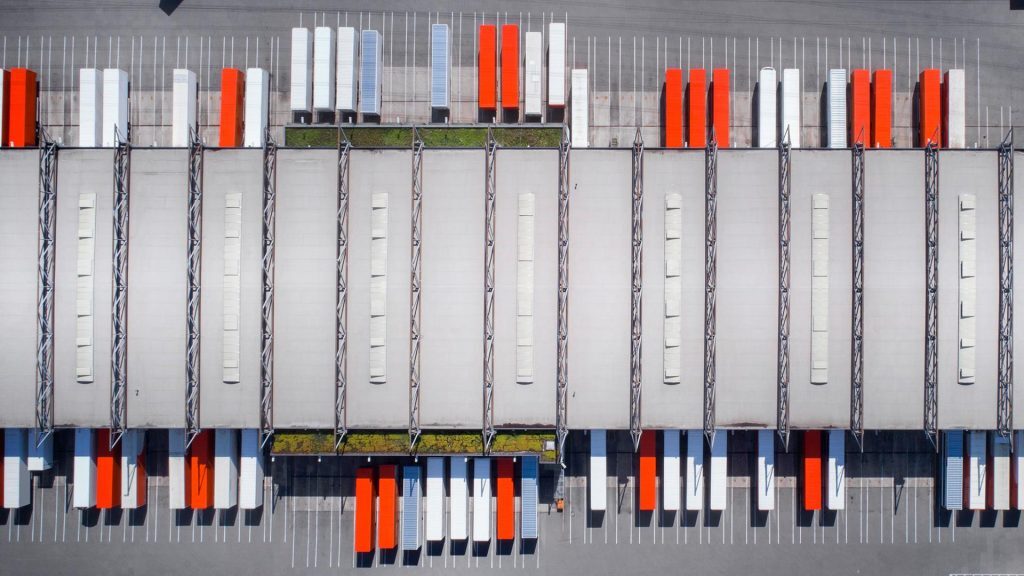 This aerial image shows a large industrial logistics or distribution center with multiple loading bays and parked semi-trailers. The trailers, in red, white, and gray, are neatly aligned along both sides of the long warehouse structure.