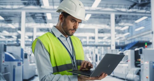 A man wearing a white hard hat and a reflective yellow safety vest is using a laptop inside a modern industrial facility or factory with bright lighting and machinery in the background.