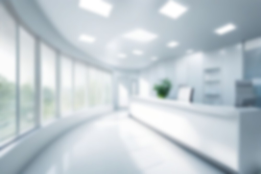 A blurred image of a bright, modern office or hospital reception area with large windows, white walls, and a sleek counter.