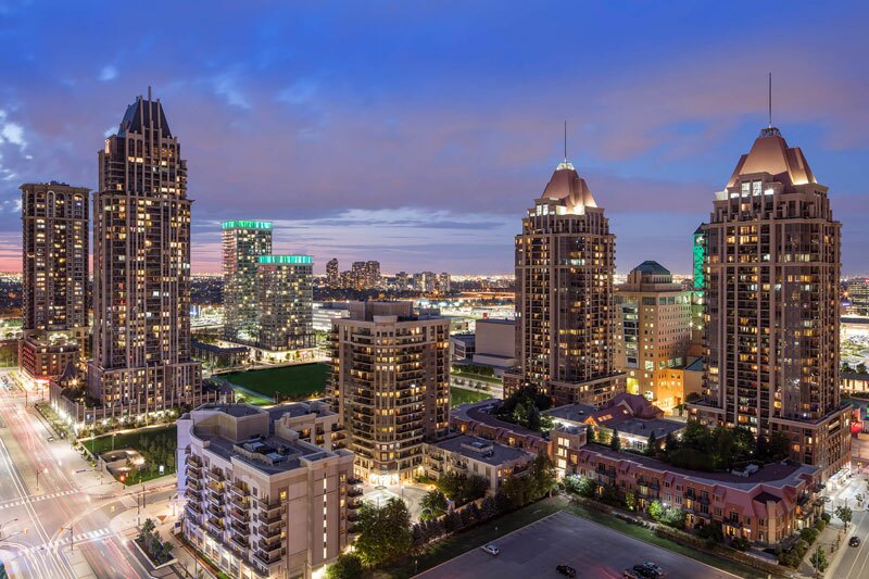 A cityscape at dusk featuring tall, illuminated residential and commercial buildings with pointed rooftops, surrounded by smaller structures, streets, and traffic lights under a blue and purple evening sky.