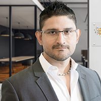 Man with short dark hair and glasses wearing a light gray suit jacket and open-collared white shirt, standing in a modern office environment.