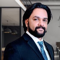 Man with medium-length dark hair and a beard wearing a dark suit, white shirt, and striped tie, posing in a modern office setting.