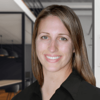A woman with straight light brown hair and a black collared shirt smiles in a modern office setting; the background shows glass walls and hanging lights, giving a professional environment impression.