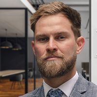 A man with short hair and a beard wearing a gray checkered suit, white shirt, and patterned tie poses in a modern office setting with glass walls and pendant lighting in the background.