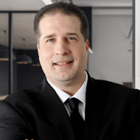 Professional man with short dark hair smiling, dressed in a black suit, white shirt, and black tie; standing in a modern office environment with glass walls and pendant lighting in the background.