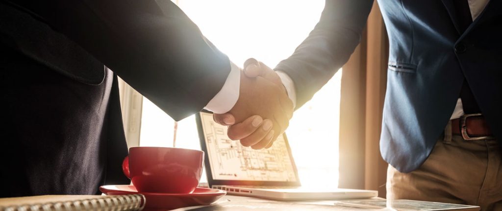 A close-up view shows two people in business attire shaking hands across a desk with a laptop, documents, and a red coffee cup. The warm lighting suggests a successful meeting or business agreement.