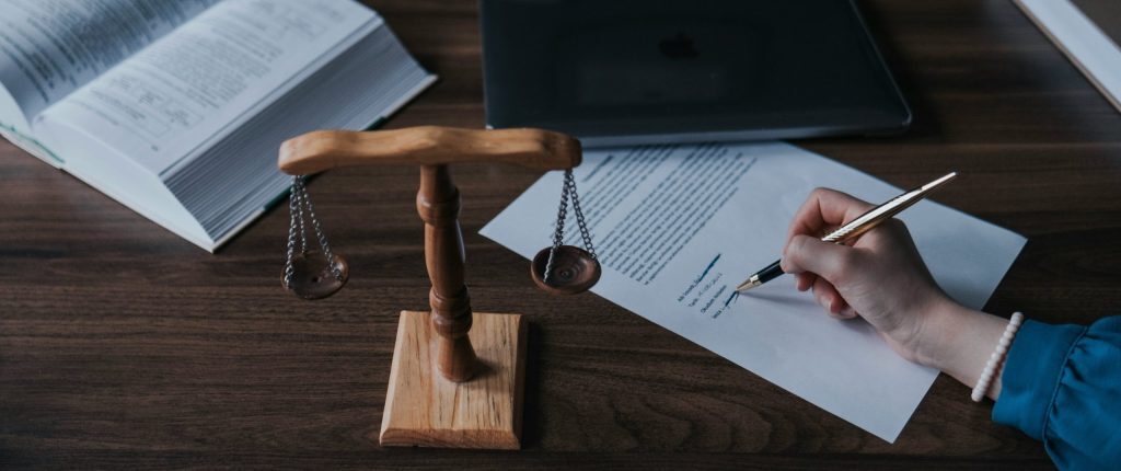 Wooden balance scale on desk beside open book and laptop – person writing or signing a document with a pen; scene symbolizes law, justice, or legal documentation.