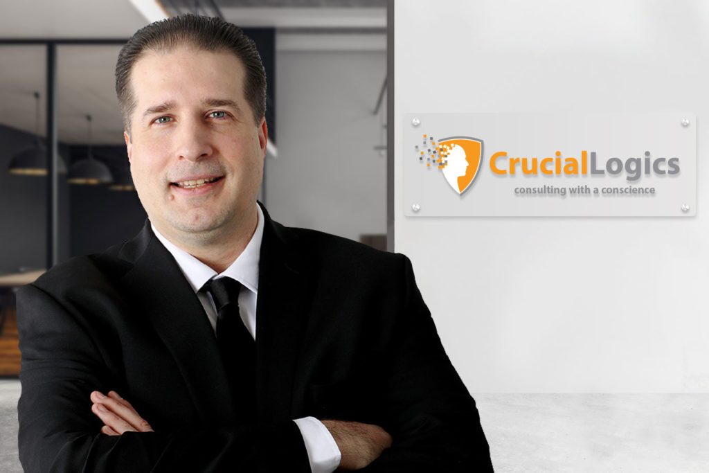 Professional man with short dark hair smiling confidently, dressed in a black suit, white shirt, and black tie; standing in a modern office beside a wall-mounted CrucialLogics logo sign that reads consulting with a conscience.