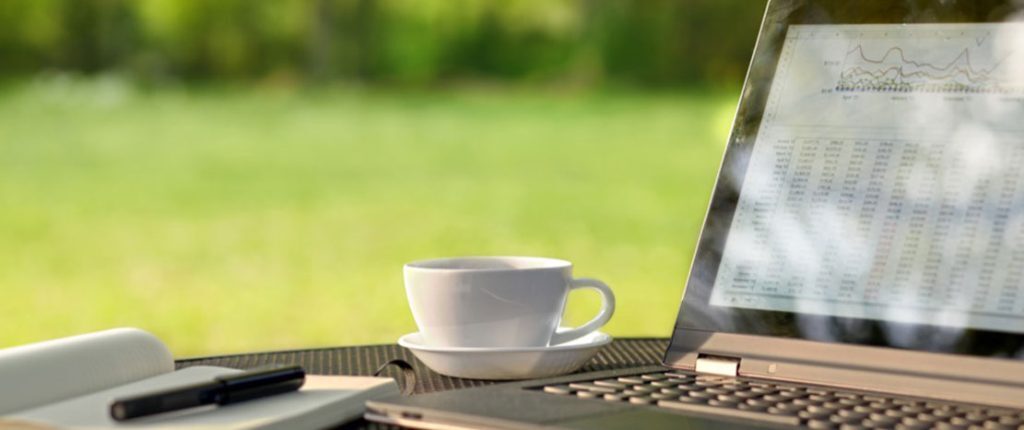 A laptop displaying charts and data sits on an outdoor table beside a white coffee cup, notebook, and pen. The background shows a blurred green lawn, suggesting remote work or a peaceful workspace setting.