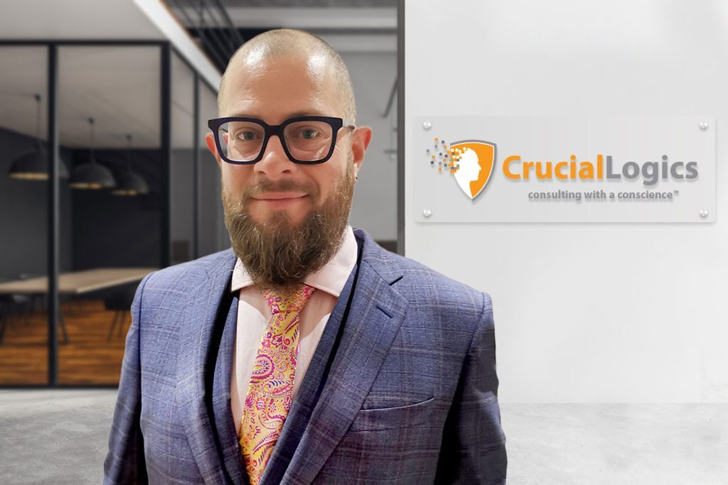 Man wearing glasses, a blue suit, and a colorful paisley tie, standing in an office environment with a CrucialLogics sign on the wall behind him.