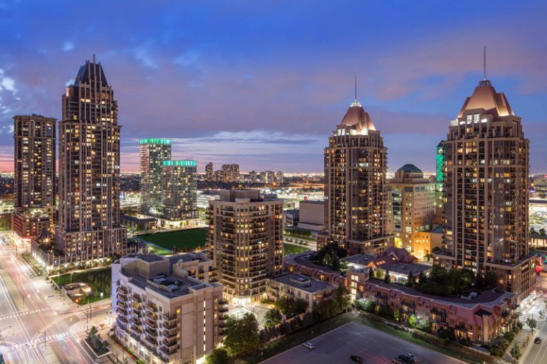 A cityscape at dusk featuring tall, illuminated residential and commercial buildings with pointed rooftops, surrounded by smaller structures, streets, and traffic lights under a blue and purple evening sky.