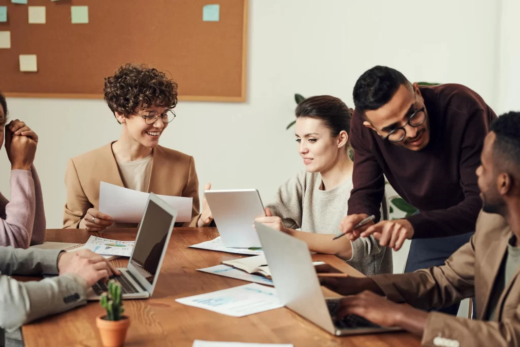A group of professionals gathered around a conference table, collaborating and discussing documents and laptops in a bright office setting.
