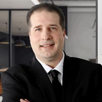 Person wearing a black suit and tie with a white shirt – smiling with arms crossed in a modern office environment; blurred glass-walled meeting area visible in background.