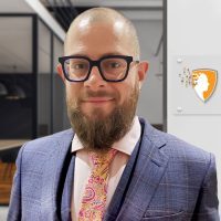 Person with a beard and glasses wearing a blue suit, pink shirt, and colorful patterned tie – standing in a modern office interior; wall plaque beside them displays an orange and gray shield logo featuring a human head silhouette with dispersing squares.
