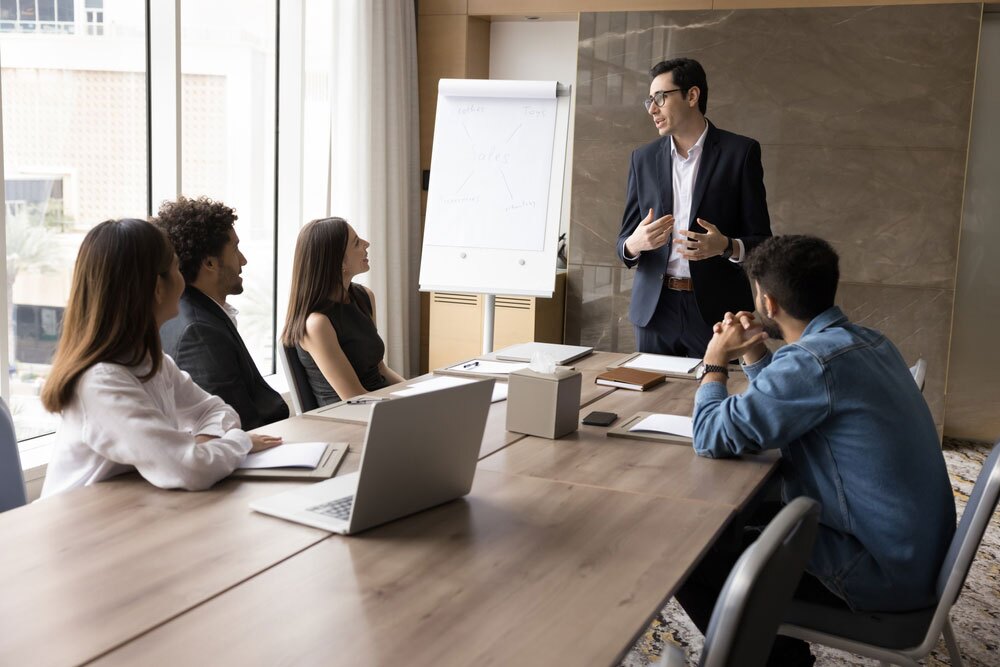 Group of professionals in a meeting room – one person standing and presenting beside a flip chart while others sit at a conference table with laptops and notebooks; scene depicts teamwork, strategy discussion, or business presentation.