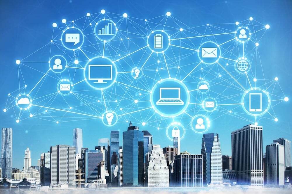 A city skyline overlaid with glowing interconnected digital icons representing devices, communication, and data exchange; symbolizing cloud computing, smart cities, and global digital networks.
