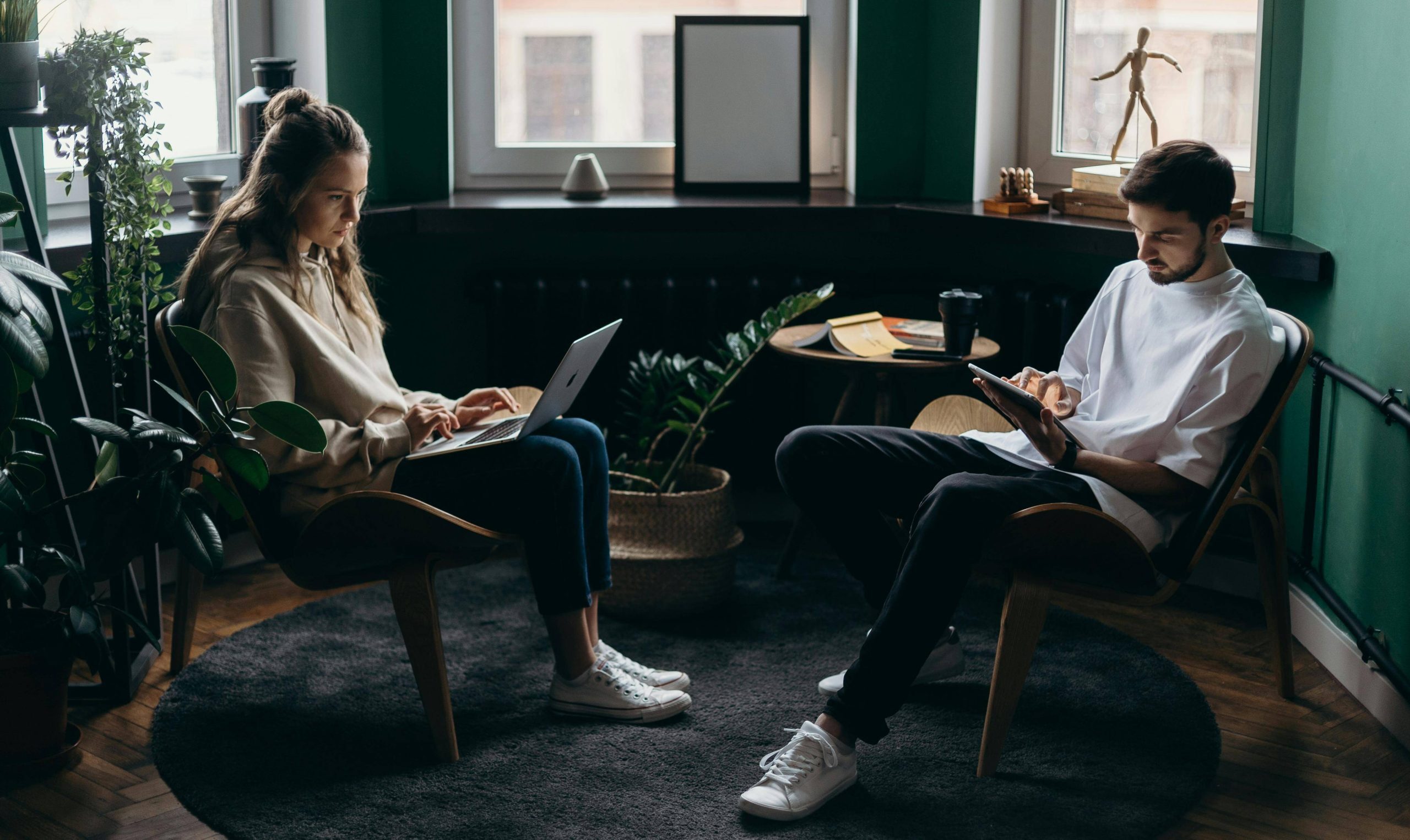 Two people sitting across from each other in a cozy, dimly lit room with green walls, working on a laptop and a tablet, surrounded by plants and modern decor.