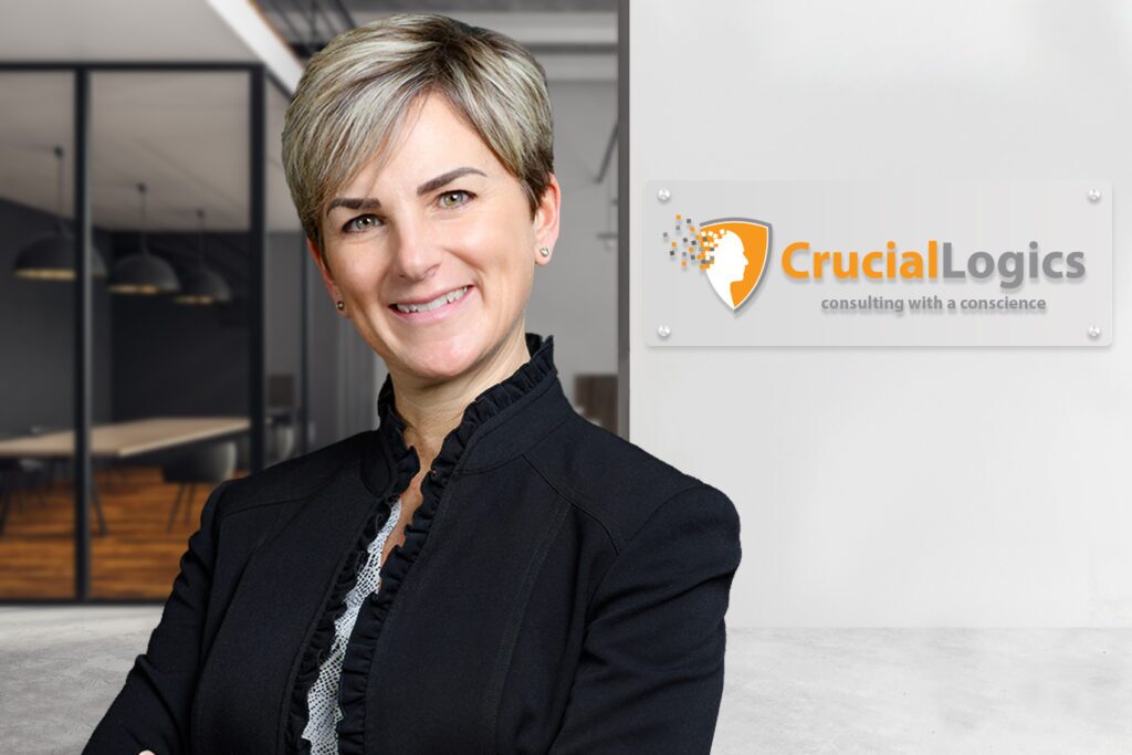 A woman in a black blazer stands confidently in a modern office setting; the Crucial Logics logo with the tagline consulting with a conscience is displayed on the wall behind her.