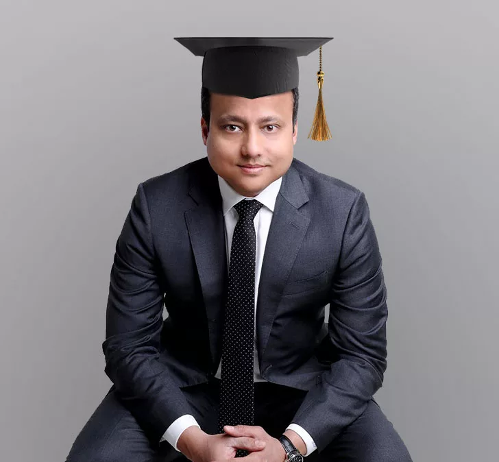 A man wearing a dark suit and tie with a graduation cap and tassel, sitting and facing the camera against a plain gray background, symbolizing achievement or professional certification.
