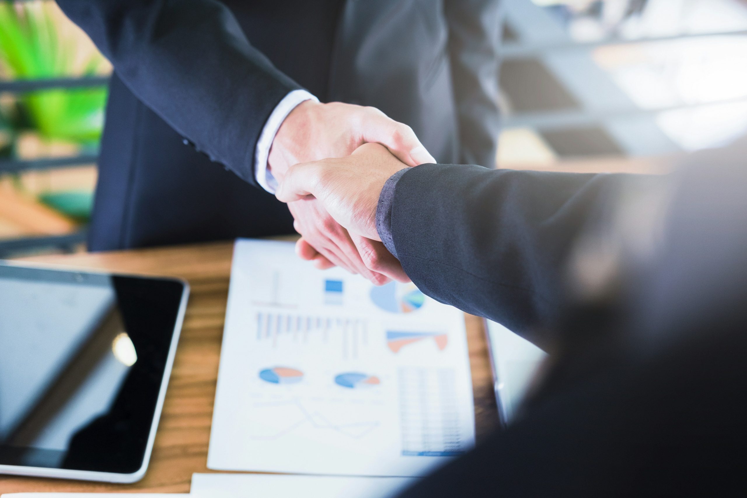 Close-up of two business professionals shaking hands over a desk with documents displaying charts and graphs, symbolizing partnership, agreement, or successful collaboration in a corporate setting.