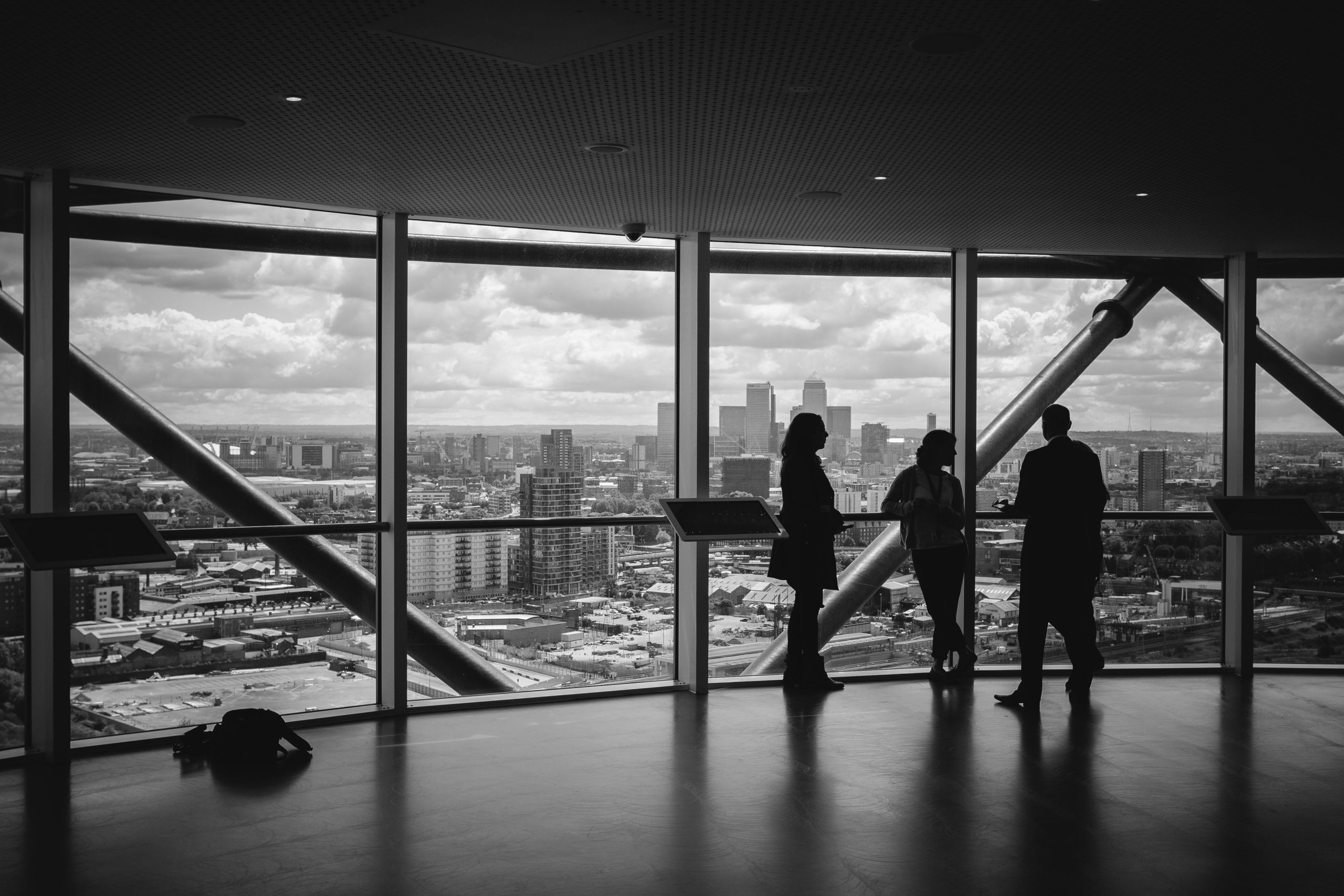 Black and white photo showing three people standing and conversing near large floor-to-ceiling windows in a high-rise building. The windows overlook a cityscape with skyscrapers and clouds in the distance, creating a professional and contemplative atmosphere.