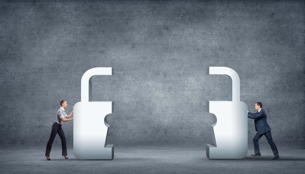 Conceptual image showing a woman and a man pushing two large metallic padlock halves toward each other against a gray textured background. The scene symbolizes collaboration, security integration, or teamwork in building a unified protection system.