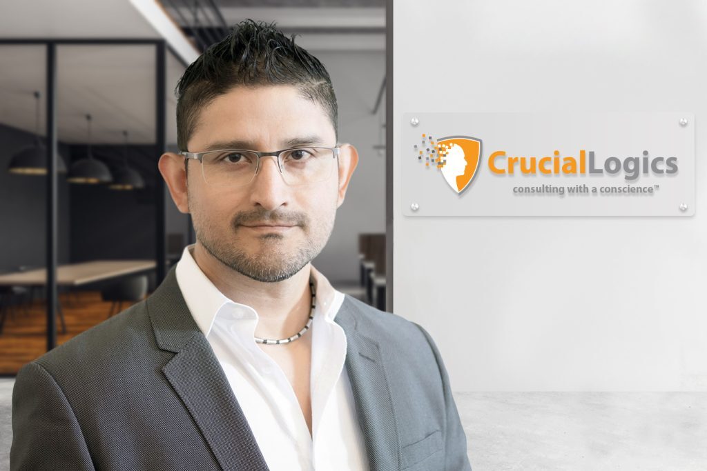 Man wearing glasses, a gray suit jacket, and an open-collared white shirt, standing in an office near a wall with the CrucialLogics logo and tagline consulting with a conscience.