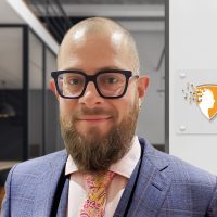 Man with glasses and a beard wearing a suit and tie, standing indoors near a wall sign displaying the CrucialLogics logo.