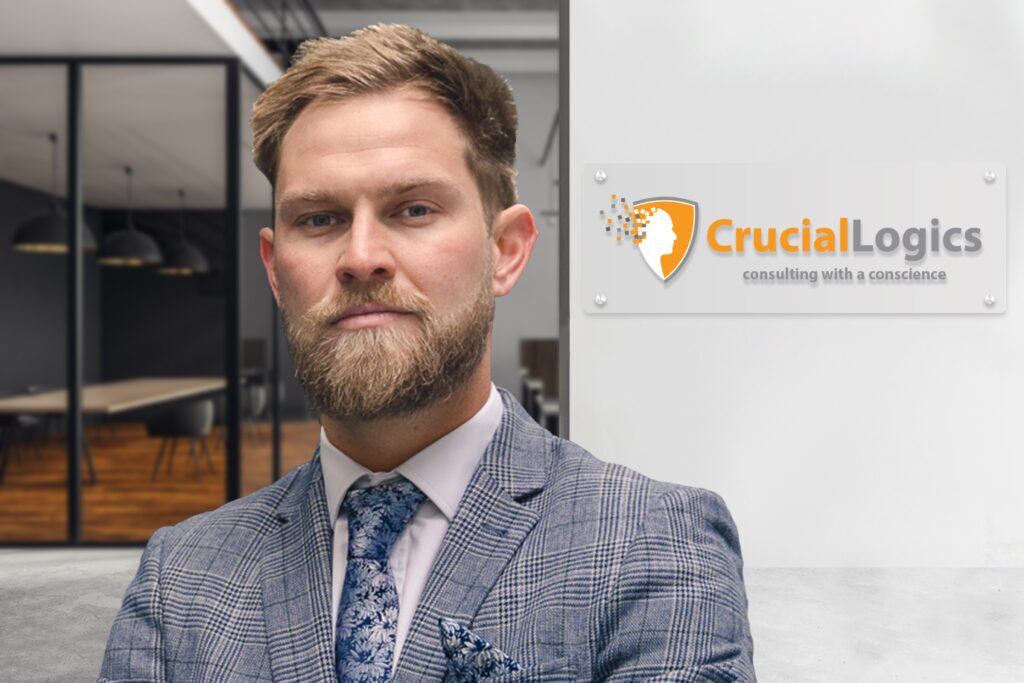 A man wearing a gray checkered suit, patterned tie, and light blue shirt stands confidently in a modern office; the Crucial Logics logo with the tagline consulting with a conscience is displayed on the wall behind him.