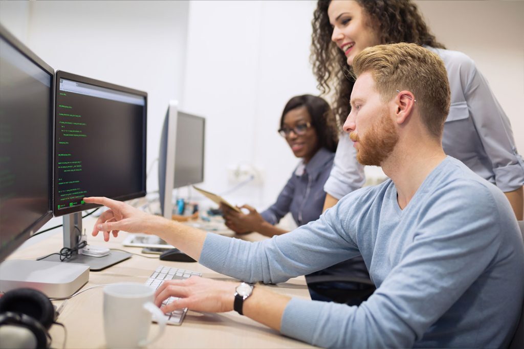 Team of professionals collaborating in a modern office – one person pointing at a computer screen displaying lines of green code while others observe and discuss; scene represents teamwork, software development, or IT problem-solving.