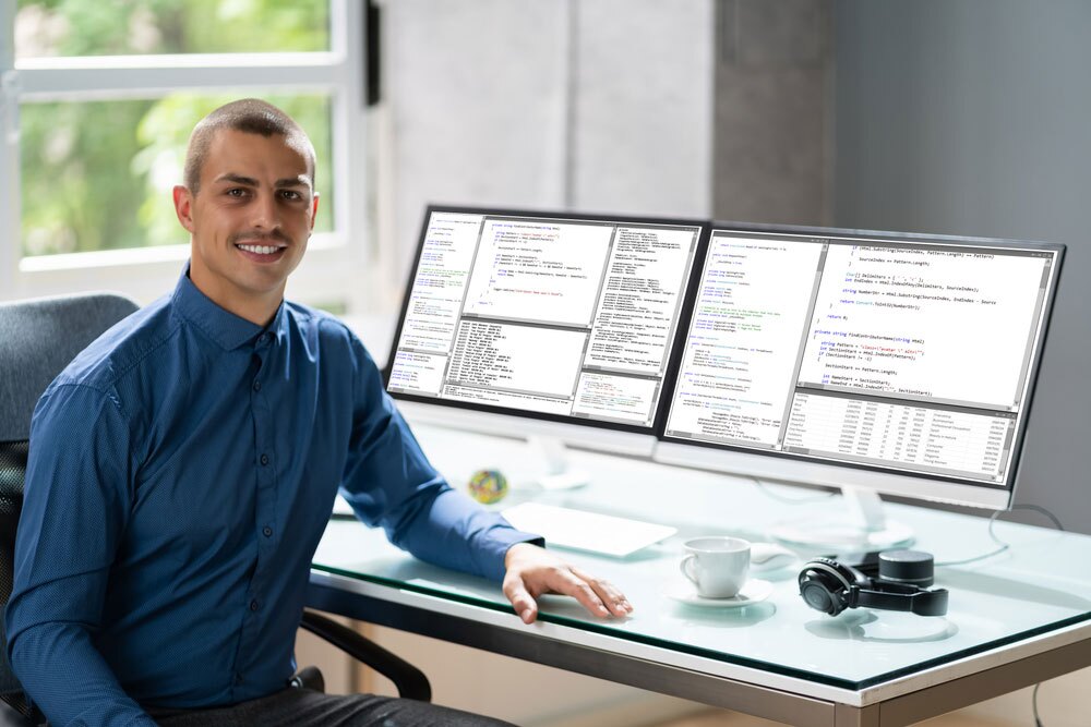 Person sitting at a desk with dual monitors displaying lines of computer code – smiling and dressed in a blue shirt; desk includes a coffee cup, headphones, and a keyboard; bright modern workspace with natural light through a window.