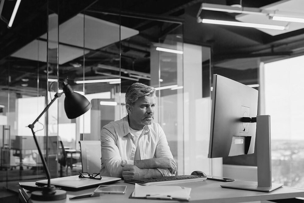 Black-and-white image of a person sitting at a desk in a modern office – focused on a computer monitor with a desk lamp, keyboard, and glasses nearby; glass walls and ceiling lights visible in the background, conveying a professional workspace.