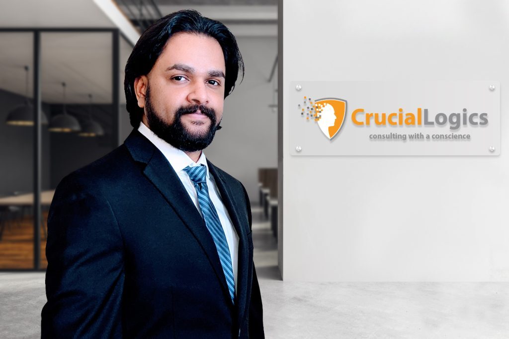 Man in a dark suit and striped tie standing in an office near a wall displaying the CrucialLogics logo and tagline consulting with a conscience.