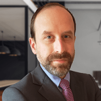 Professional man with a short beard and receding hairline wearing a dark suit, light blue shirt, and pink patterned tie; posed confidently in a modern office environment with glass partitions and soft lighting.