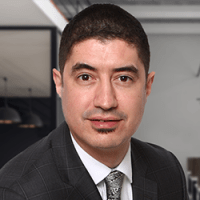 Professional man with short dark hair wearing a gray checkered suit, white shirt, and patterned tie; posed confidently in a modern office setting with neutral tones and glass partitions in the background.