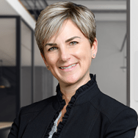 Professional woman with short blonde hair smiling confidently, dressed in a black blazer and standing in a modern office environment with glass walls and soft lighting.